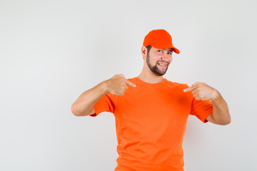 Delivery man in orange t-shirt, cap pointing at himself and looking proud , front view.